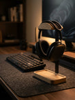Black over-ear headphones displayed on a wooden stand on a desk, with a keyboard and monitor in a dimly lit home office setting.