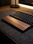 A close-up of a black mechanical keyboard resting on a dark gray felt desk mat, with a wooden wrist rest in front of it and a blurred desk environment in the background.